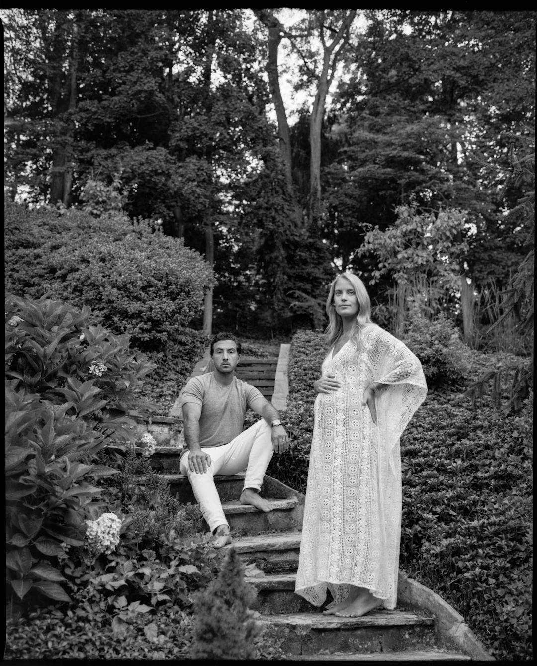 black and white medium format maternity portrait of woman in white dress holding belly with man seated on stone stairs